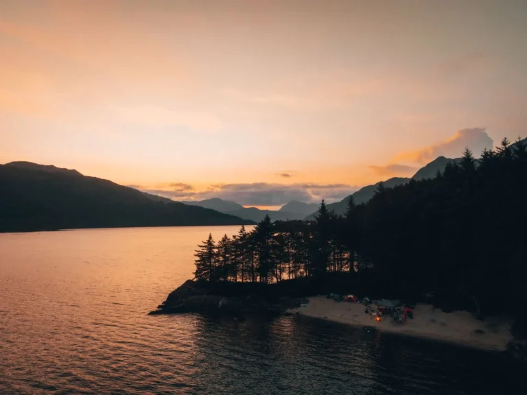 Camping au bord d’un lac au coucher du soleil, entouré d’arbres et de montagnes